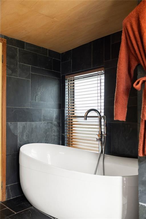 Bathroom with a white freestanding tub, dark tile, wood ceiling, and window blinds.