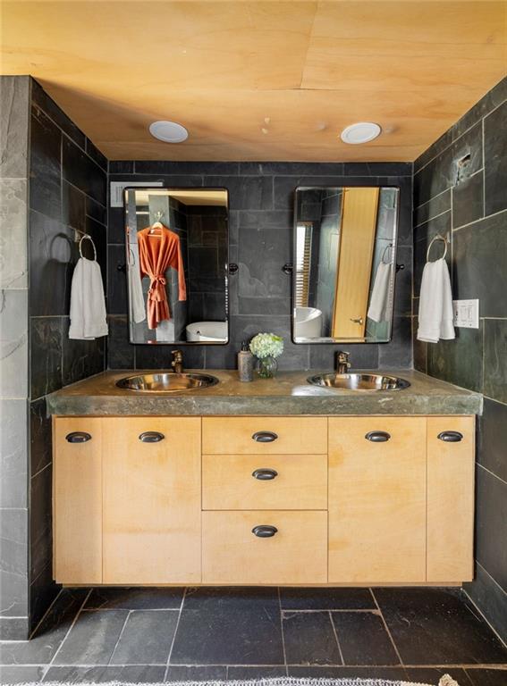 Bathroom with a double-sink vanity, light wood cabinets, dark tile, and two mirrors.