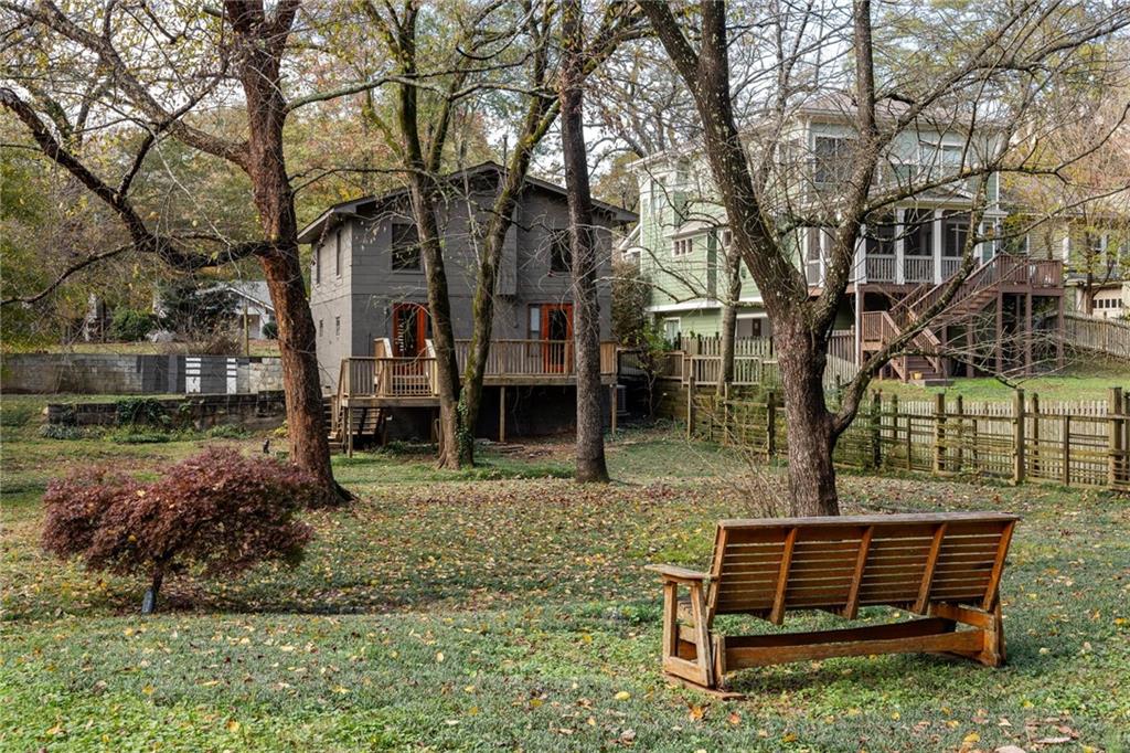 Gray house and raised deck beyond a leaf-strewn backyard, with a wooden bench beneath mature trees.