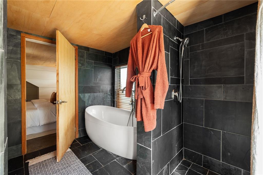 Bathroom with dark stone tile, freestanding white tub, wood ceiling, and rust-colored robe.