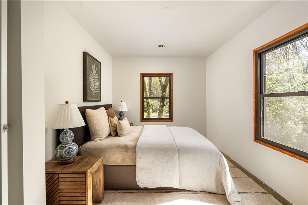 Bedroom with white and beige bedding, a wooden bedside table, and wood-trimmed windows overlooking trees.