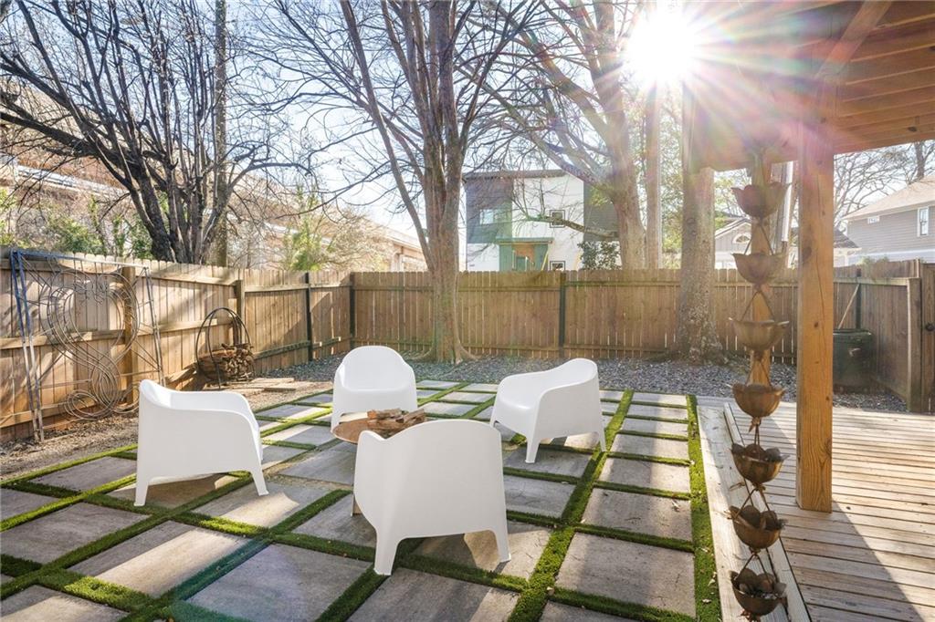 Backyard patio with white chairs, grass-lined stone pavers, and a wooden deck beneath leafless trees.