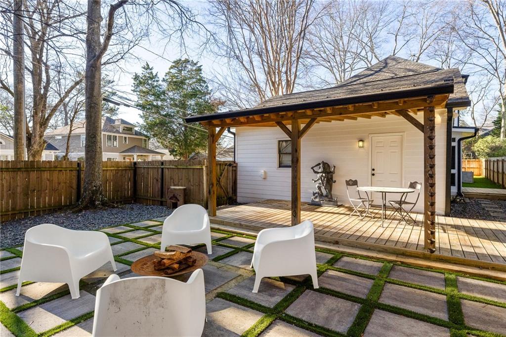 Backyard patio with a covered wooden deck, white lounge chairs, and a square-paver fire-pit area.