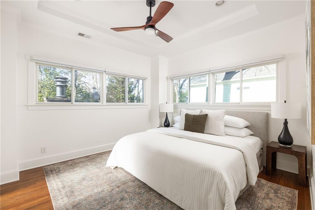 Staged bedroom with a white bed, gray headboard, two walls of windows, and a wood ceiling fan.