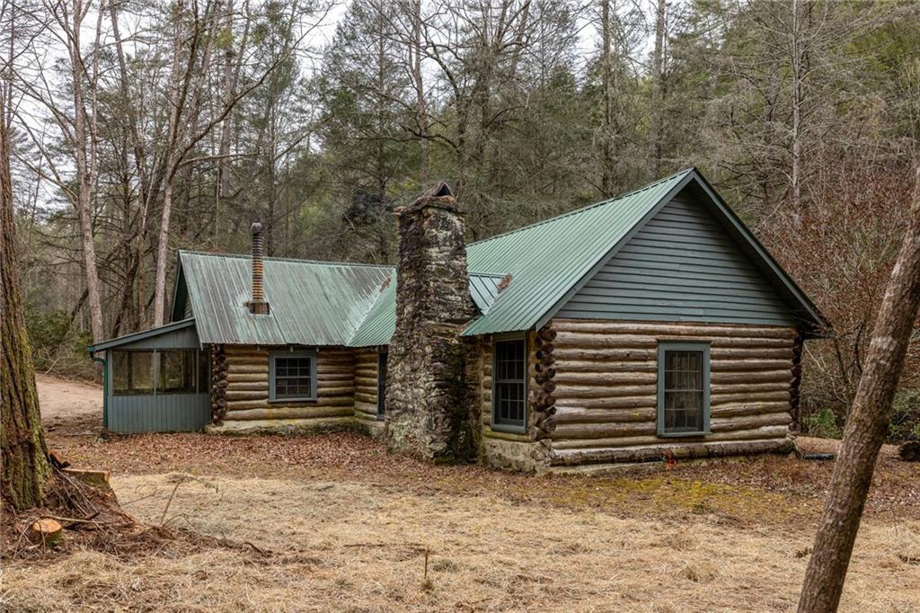 Log cabin with green metal roofs and a tall stone chimney in a wooded clearing.