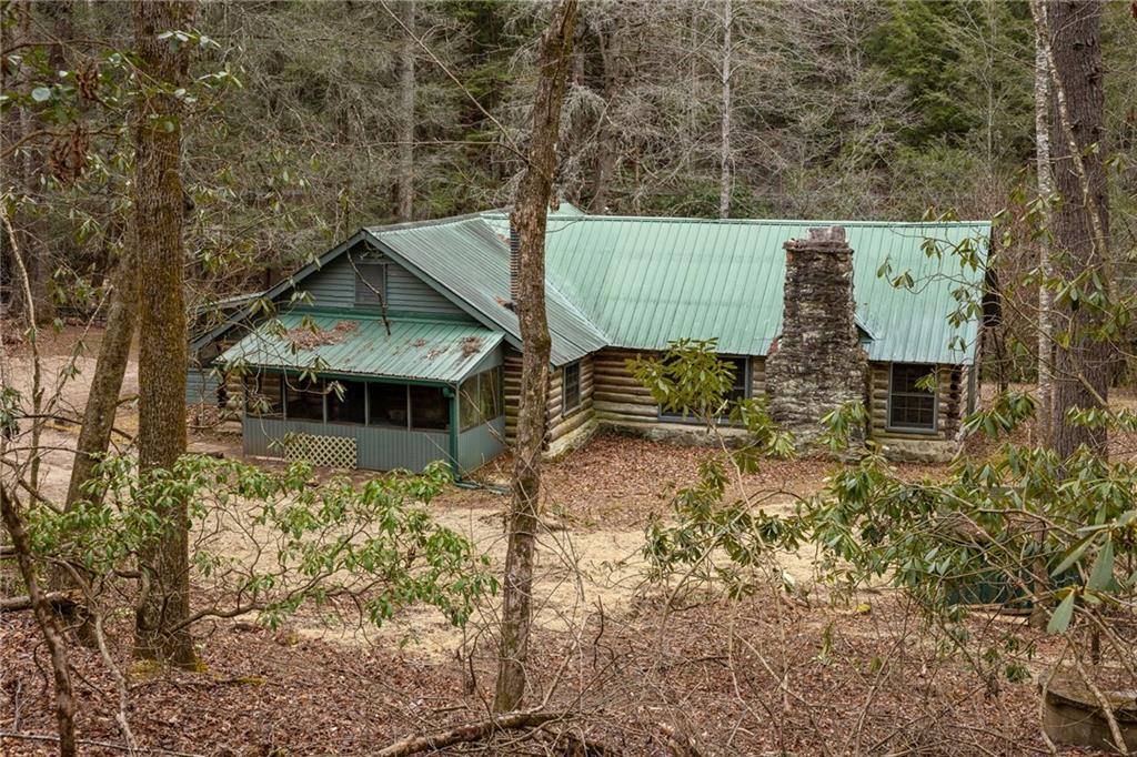 Woodland log cabin with a green metal roof, stone chimney, and covered porch.
