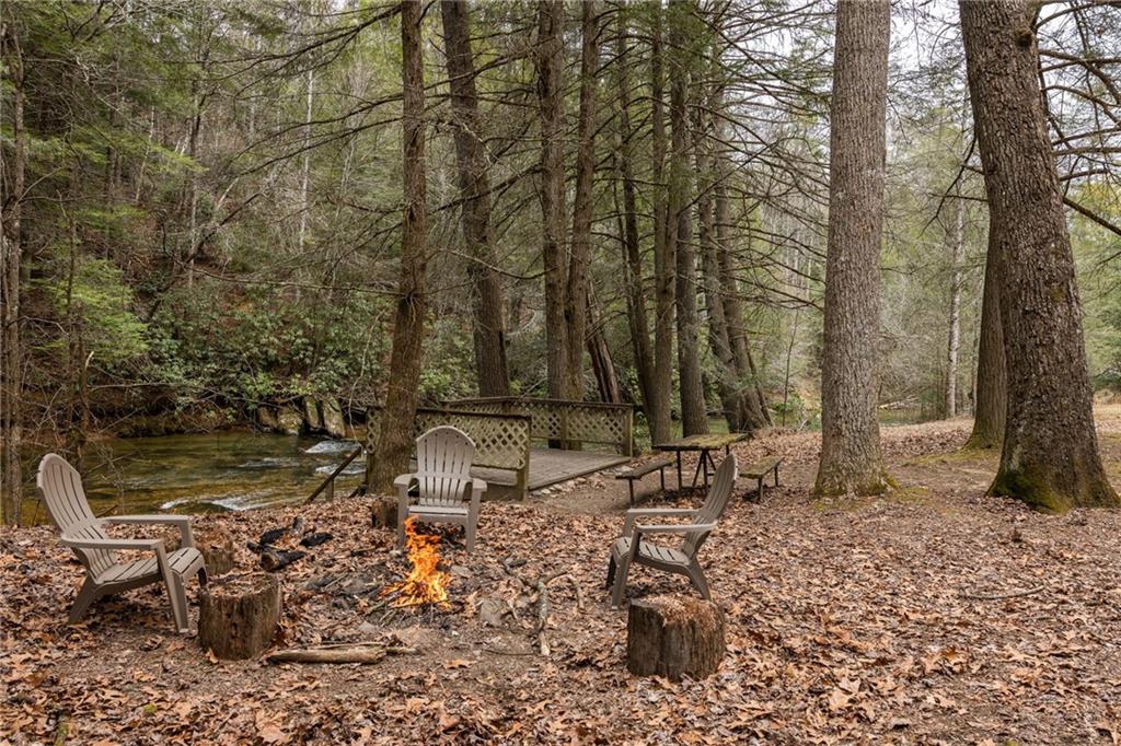 Wooded creekside seating area with Adirondack chairs around a fire pit beside a wooden footbridge.