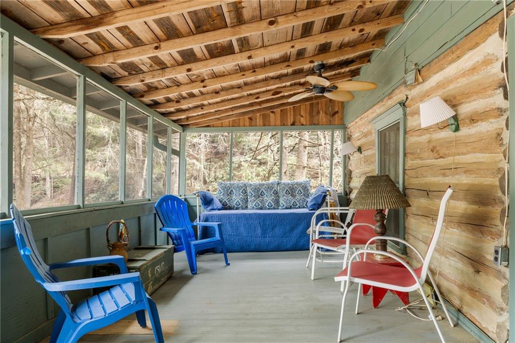 Screened porch with log walls, exposed wood beams, colorful seating, and a wooded view.