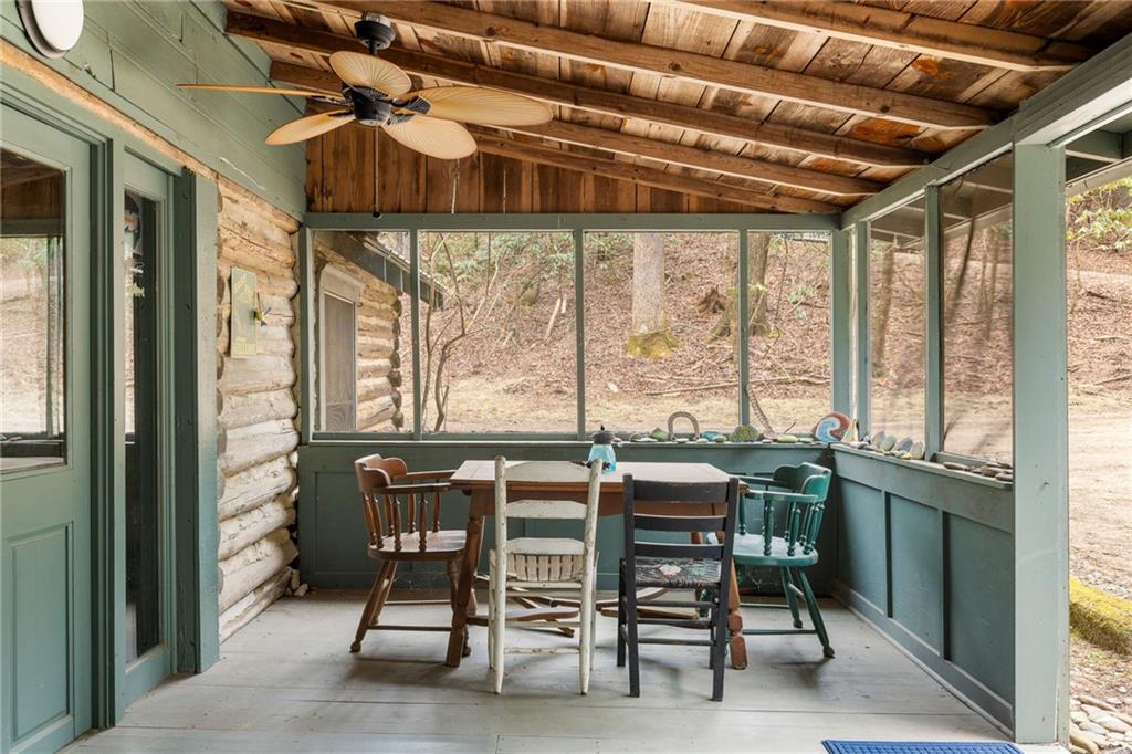 Enclosed porch with a dining table, log walls, and large windows overlooking a wooded hillside.