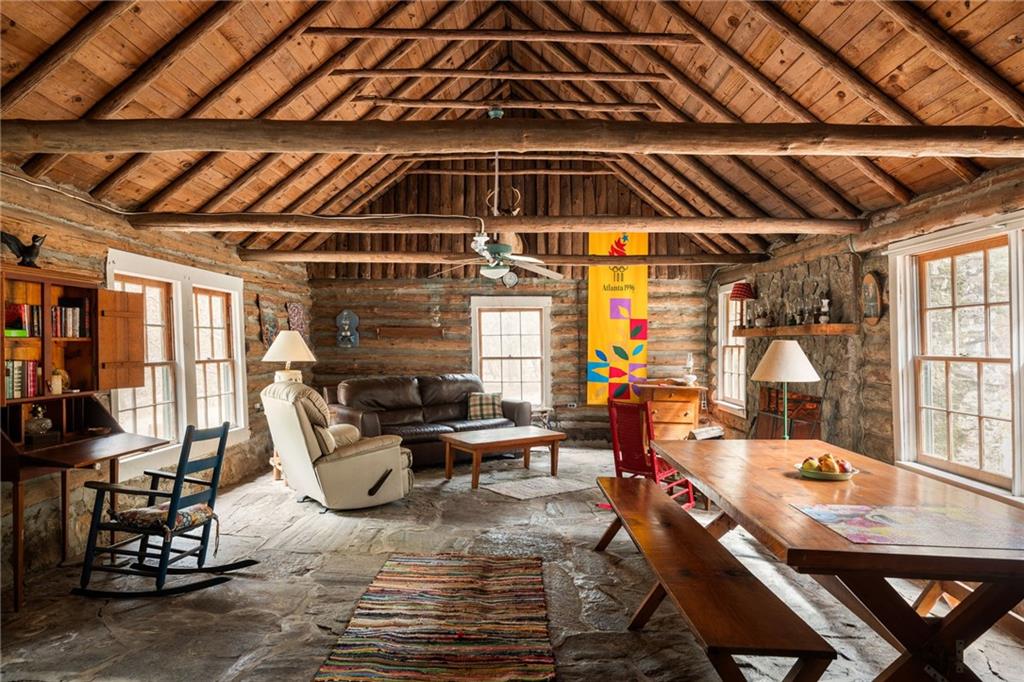 Rustic cabin living room with log walls, exposed timber ceiling, stone floor, and a long dining table.