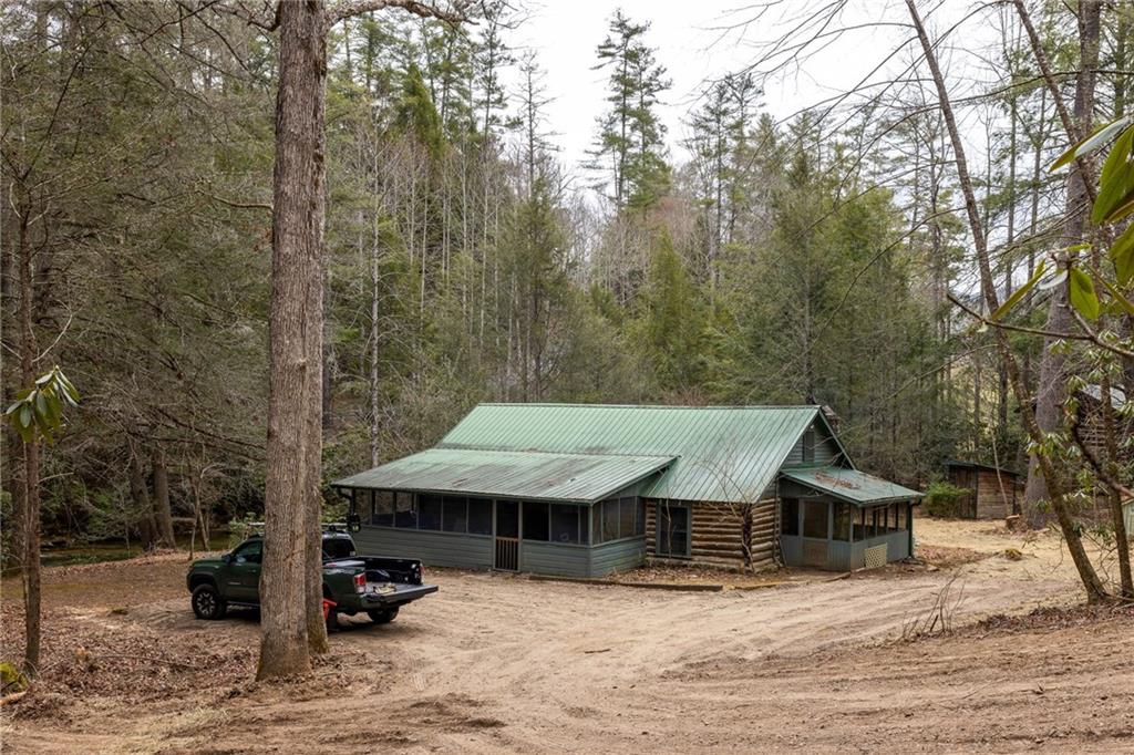 Rustic log cabin with a green metal roof sits among trees beside a dirt driveway.