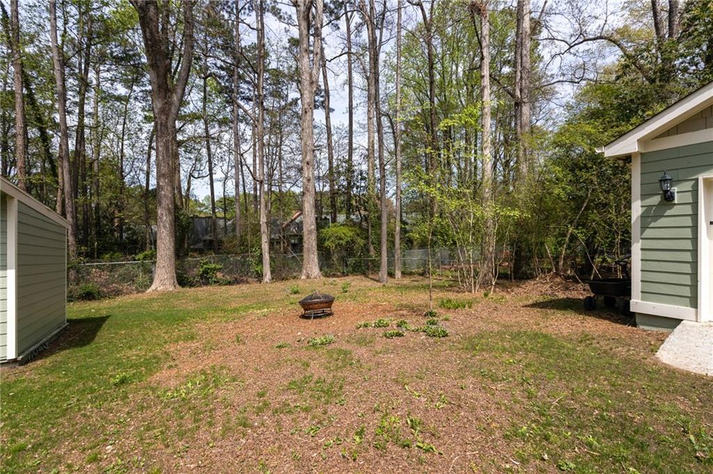Backyard with a fire pit, patchy lawn, and tall trees beyond a chain-link fence.