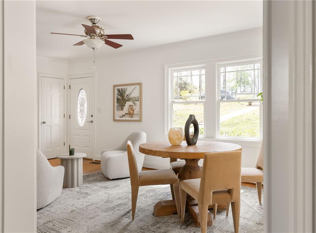 Dining area with a round wooden table, upholstered chairs, and bright windows beside a small sitting area.