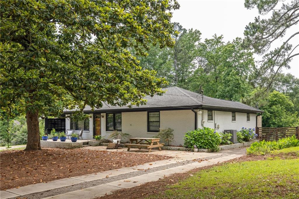 Single-story brick ranch house beneath mature trees, with a paved drive and patio furniture.