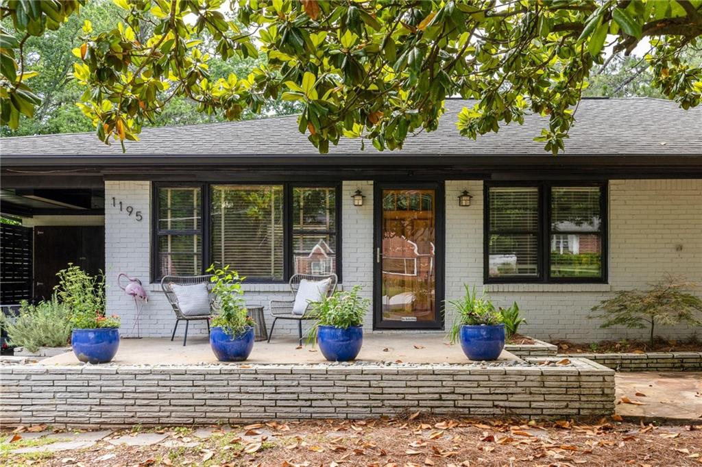 White brick house with dark-framed windows, raised patio, blue planters, and mature tree.