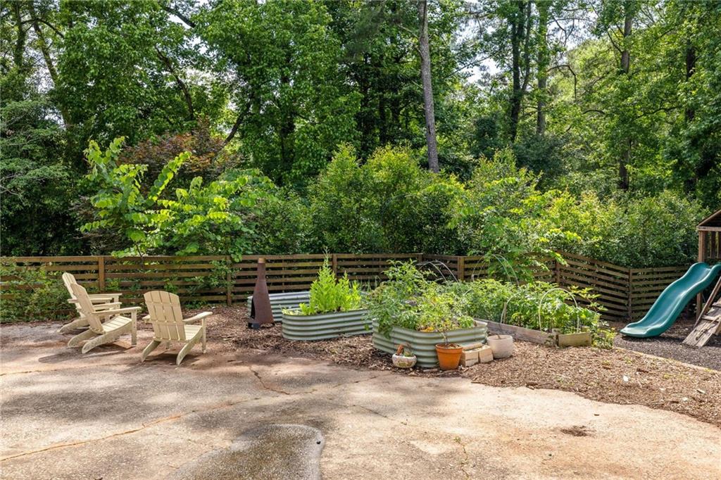 Wooded backyard patio with Adirondack chairs, raised garden beds, and a green slide.