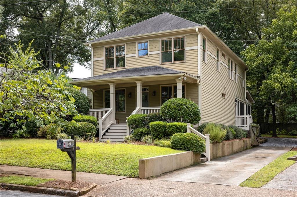 Two-story beige house with a covered porch, white railings, landscaped shrubs, and a driveway.