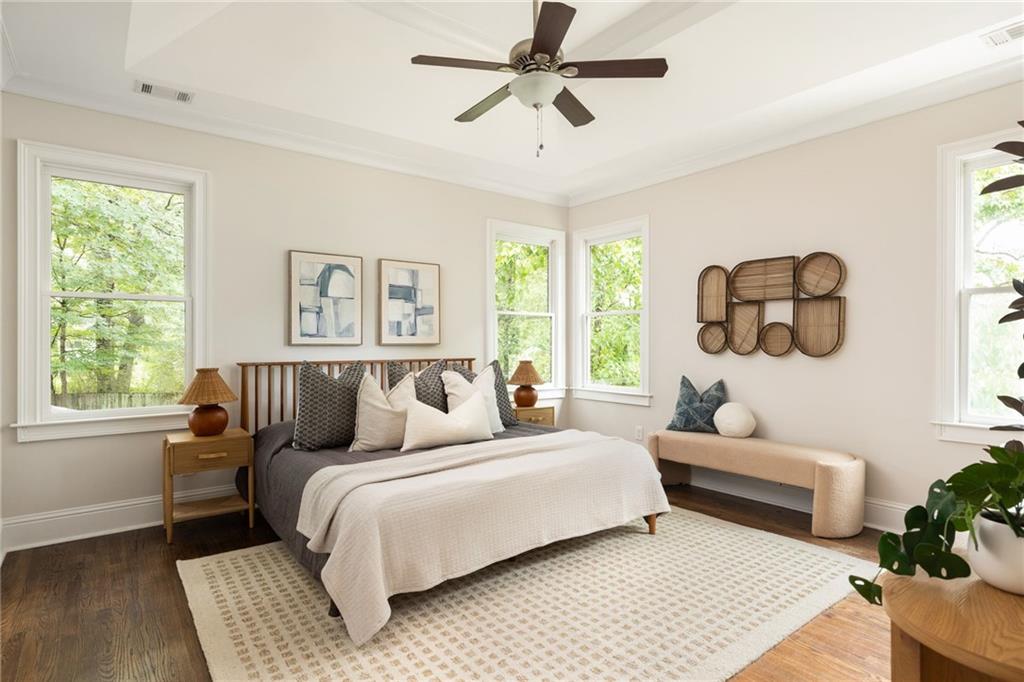 Bright bedroom with a white-covered wooden bed, cream bench, and windows overlooking leafy trees.