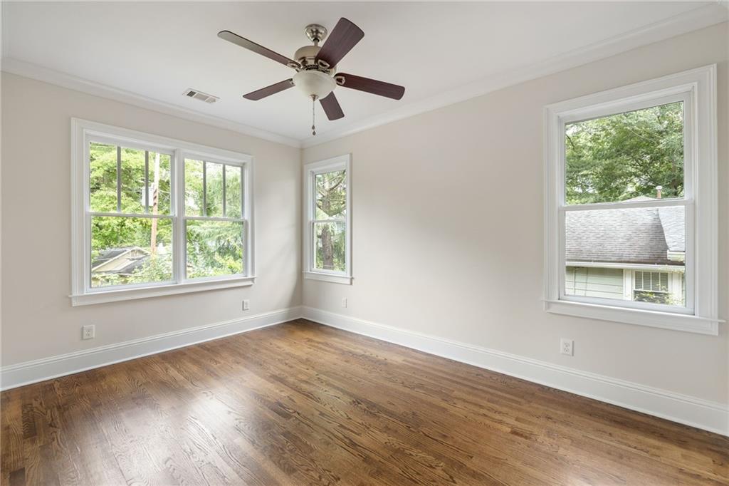 Empty room with polished hardwood floors, a ceiling fan, and windows overlooking leafy trees.
