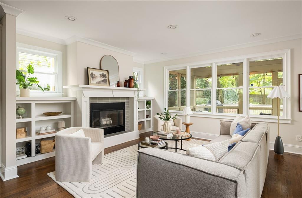 Living room with a fireplace, built-in shelves, and large windows overlooking greenery.
