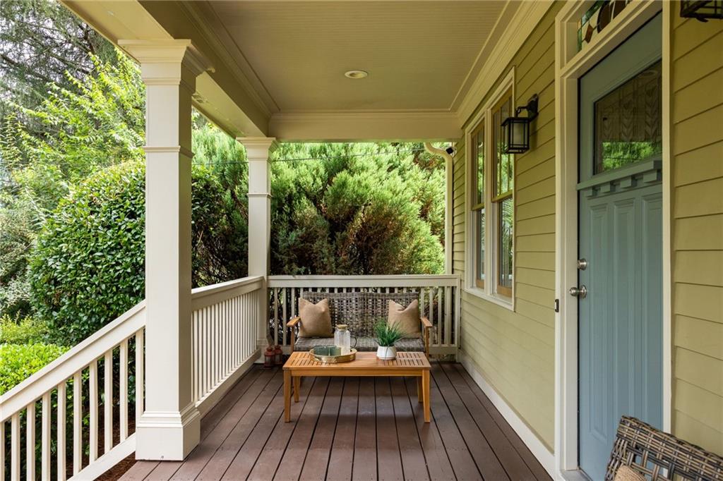 Covered front porch with a blue door, white railings, and a small seating area surrounded by greenery.