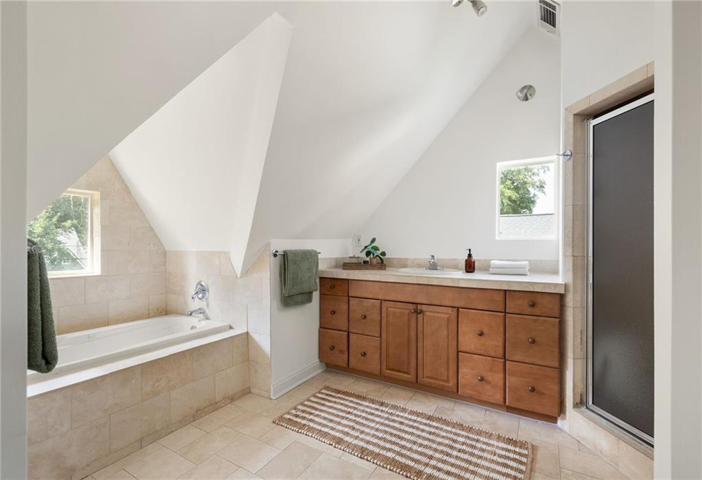 Bright bathroom with a tiled bathtub, wooden vanity, and glass-enclosed shower beneath a sloped ceiling.