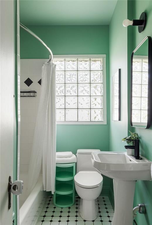 Mint-green bathroom with glass-block window, geometric tile floor, pedestal sink, and small towel shelf.