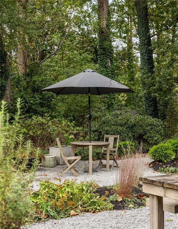 Garden patio with a round table and chairs beneath an umbrella, surrounded by trees and lush planting.