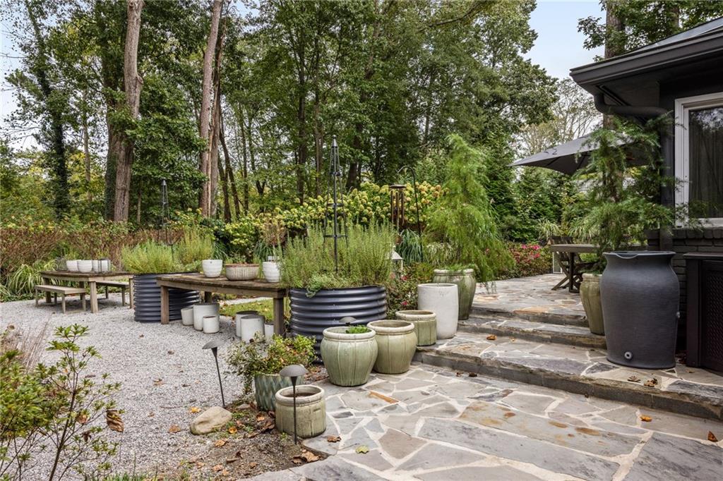 Stone patio beside a dark-sided home, surrounded by planters, raised beds, and mature greenery.