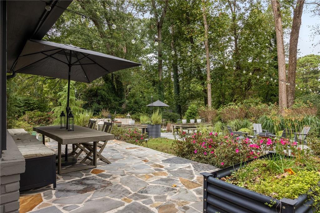 Stone patio with umbrella-shaded dining table, string lights, garden seating, and flowering shrubs.