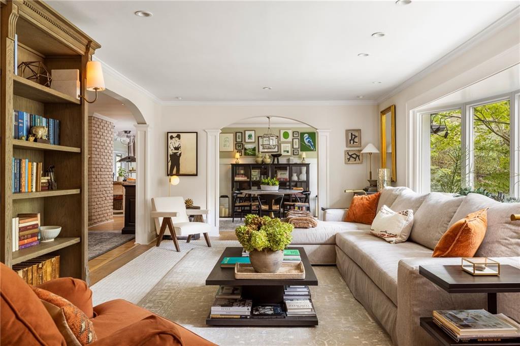 Living room with a beige sectional, orange accent chairs, built-in bookcase, and leafy views through large windows.