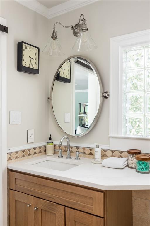 Bathroom vanity with wood cabinetry, oval mirror, and decorative tile backsplash beside a bright window.