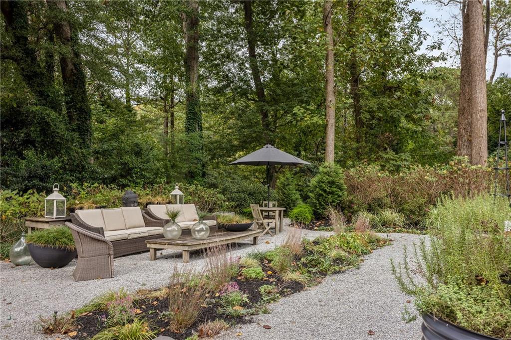 Gravel garden patio with wicker seating, a wooden coffee table, and a gray umbrella beneath mature trees.