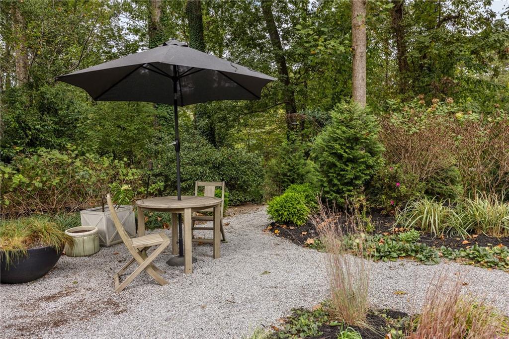 Gravel patio with a round table and chairs beneath a black umbrella, surrounded by landscaped shrubs and trees.