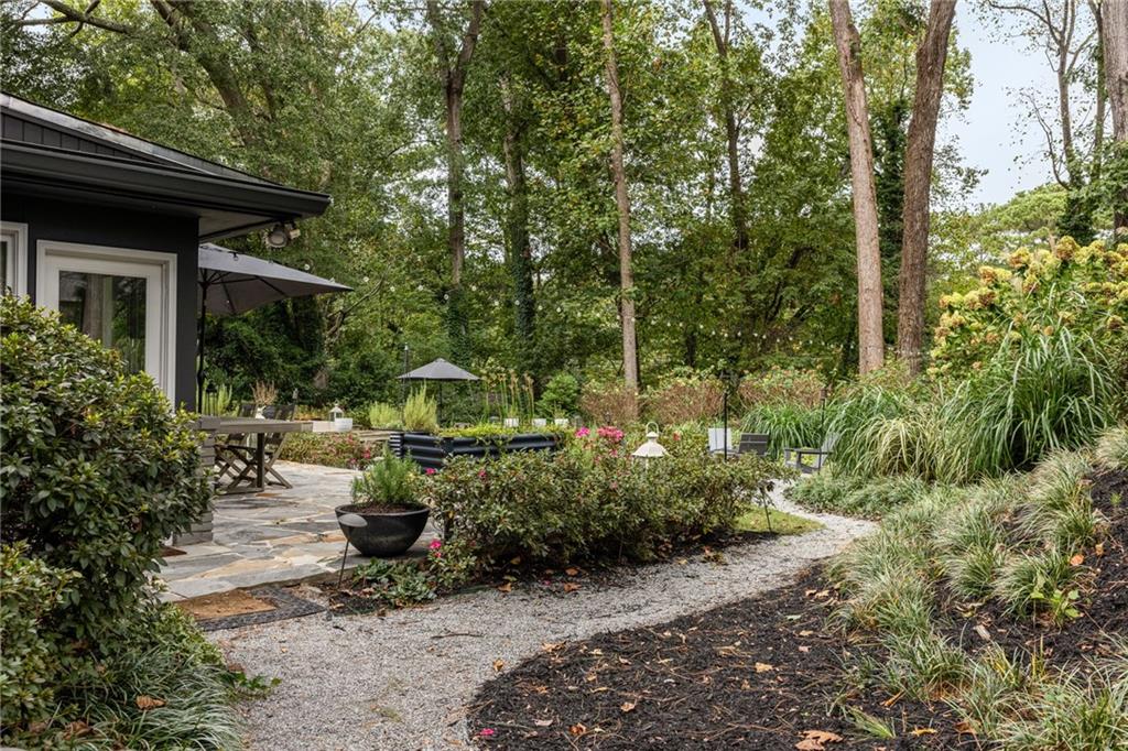 Landscaped patio with stone paving, outdoor seating, flowering shrubs, and a gravel path beneath mature trees.