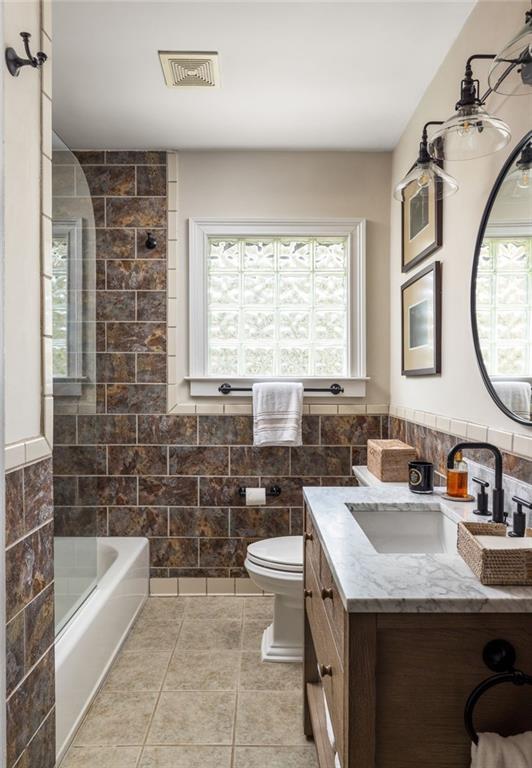 Bathroom with brown tiled walls, a glass-block window, white bathtub, and wood vanity with a marble-look counter.
