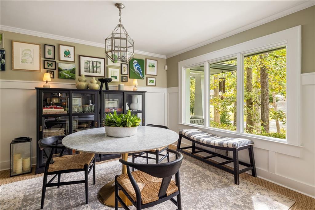 Dining room with a round table, glass-front cabinet, and broad windows overlooking leafy trees.