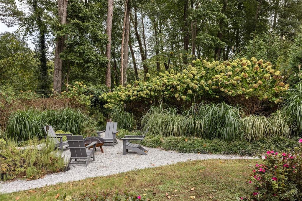 Landscaped backyard seating area with Adirondack chairs on gravel among flowering shrubs and mature trees.