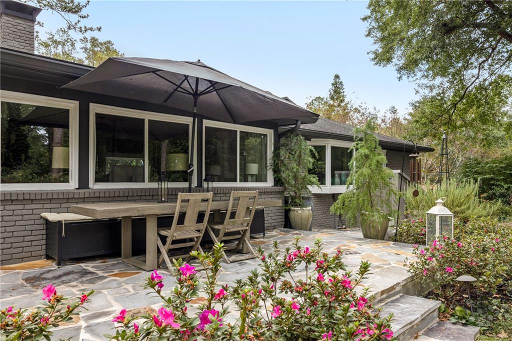 Brick home’s stone patio with an umbrella-shaded dining set and pink flowering shrubs.