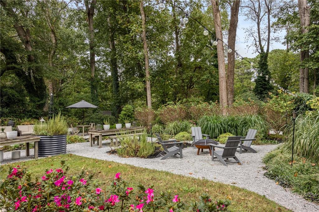 Gravel patio with lounge chairs around a fire pit beneath mature trees and string lights.