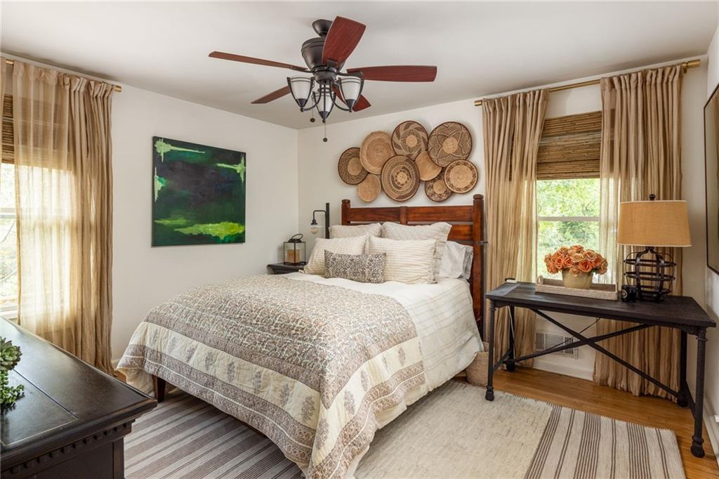 Furnished bedroom with cream patterned bedding, woven wall decor, and beige curtains framing two windows.