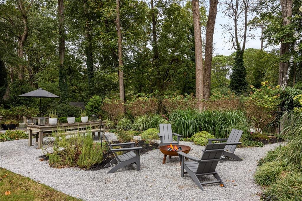 Gravel backyard patio with Adirondack chairs around a fire pit, bordered by mature trees and lush planting.