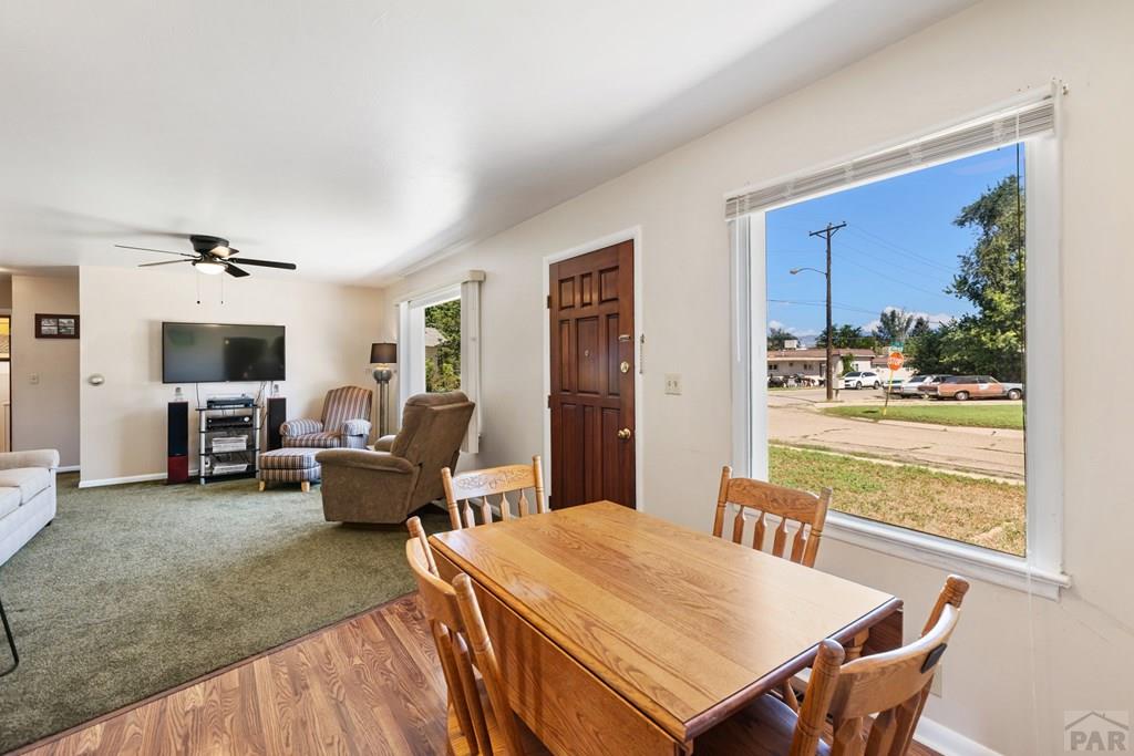 Dining area with a wooden table beside a large street-facing window, opening onto a carpeted living room.
