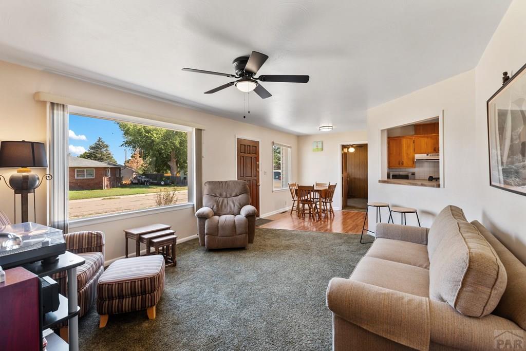 Living room with a large street-facing window, gray carpet, beige sofa, and recliner.