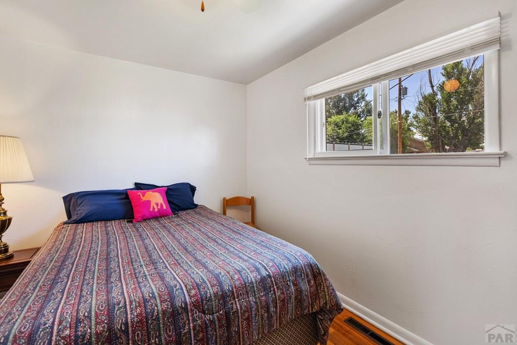 Furnished bedroom with a patterned bedspread, bright pink accent pillow, and wide window overlooking trees.