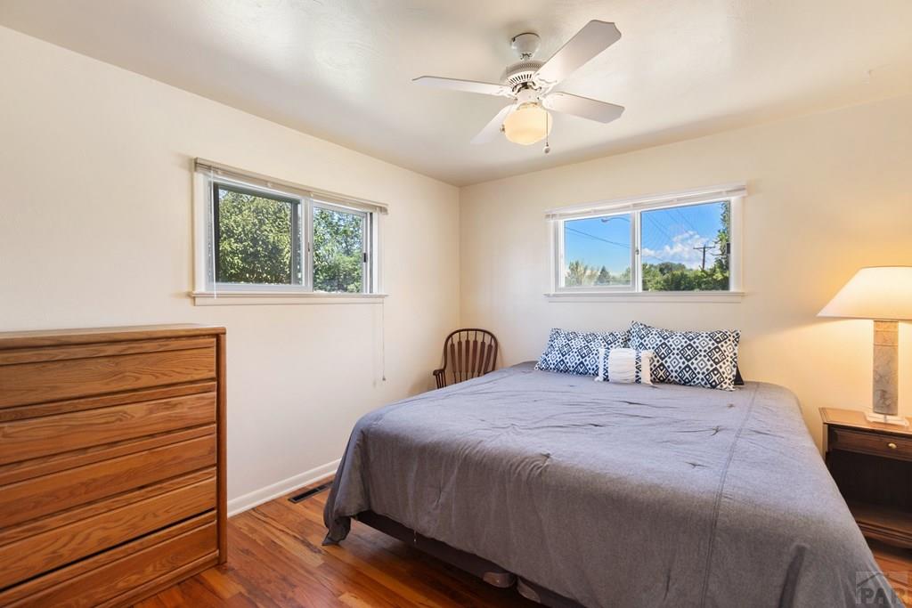 Bedroom with a gray-covered bed, wooden dresser, hardwood floors, and two windows.