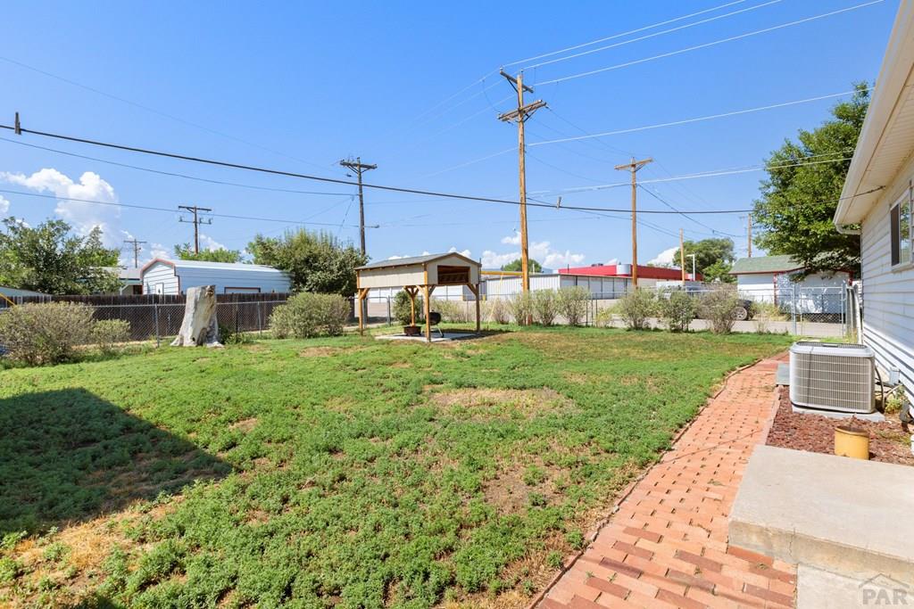 Backyard with a patchy lawn, brick walkway, wooden shelter, and chain-link fence.