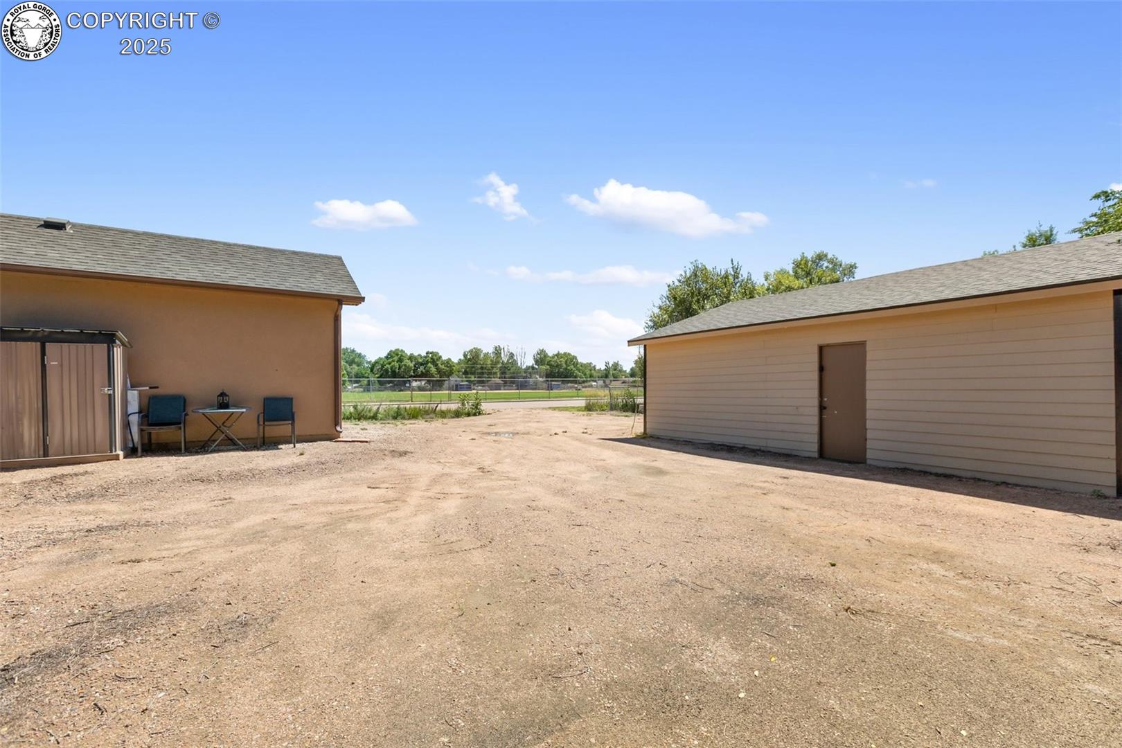Gravel yard between a tan house and detached beige garage, with patio chairs and a small table.