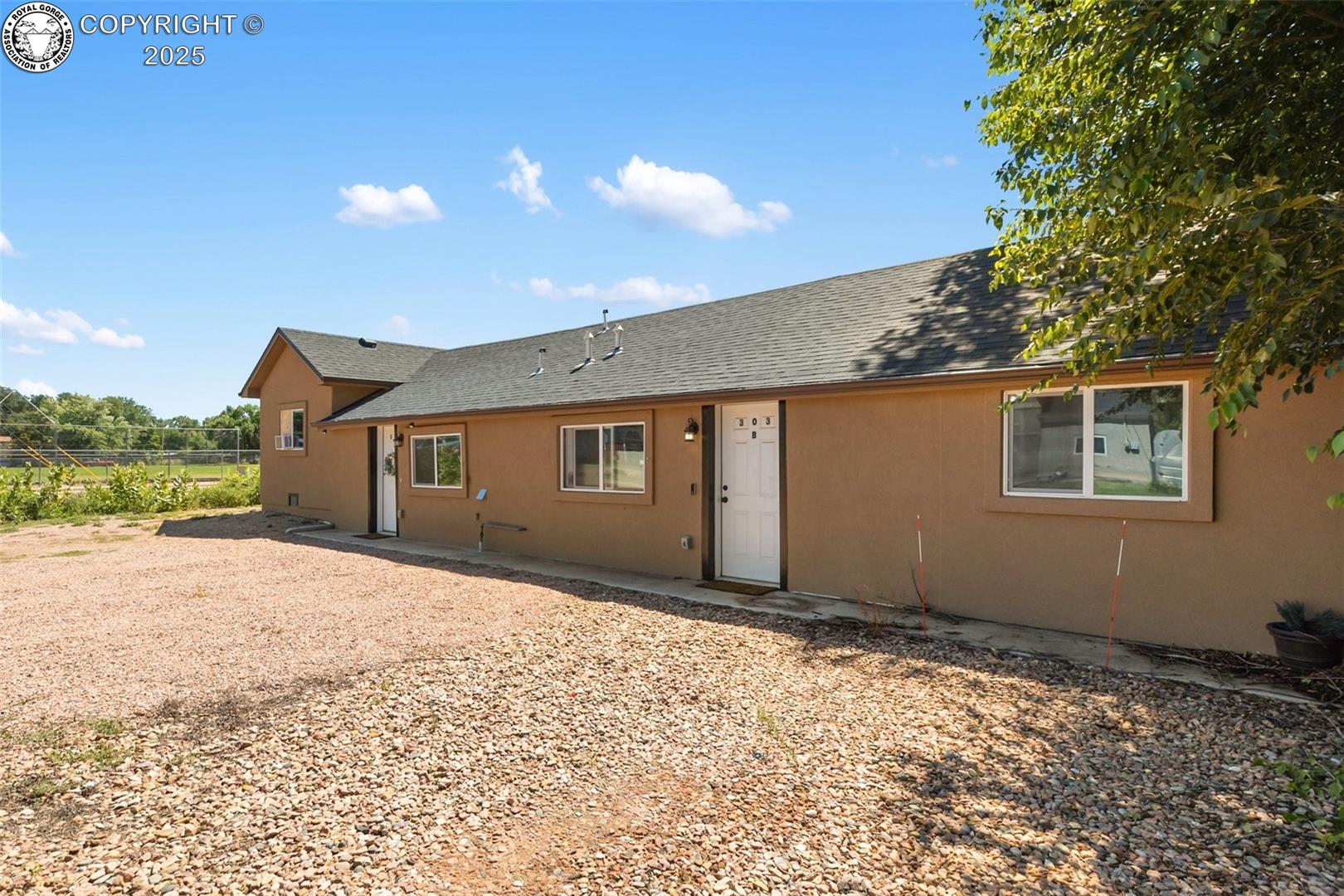 Tan stucco home with a dark shingle roof, white entry door, and broad gravel yard.