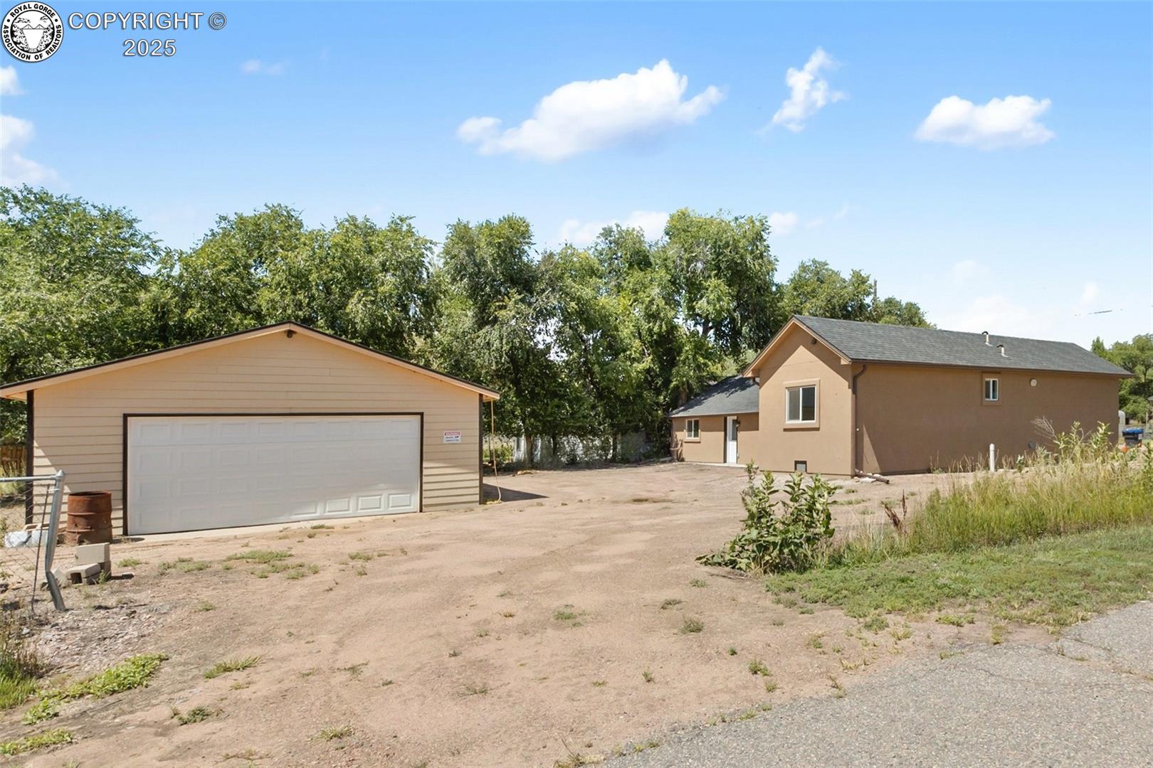 Tan single-story home and detached two-car garage beside a broad dirt driveway and mature trees.